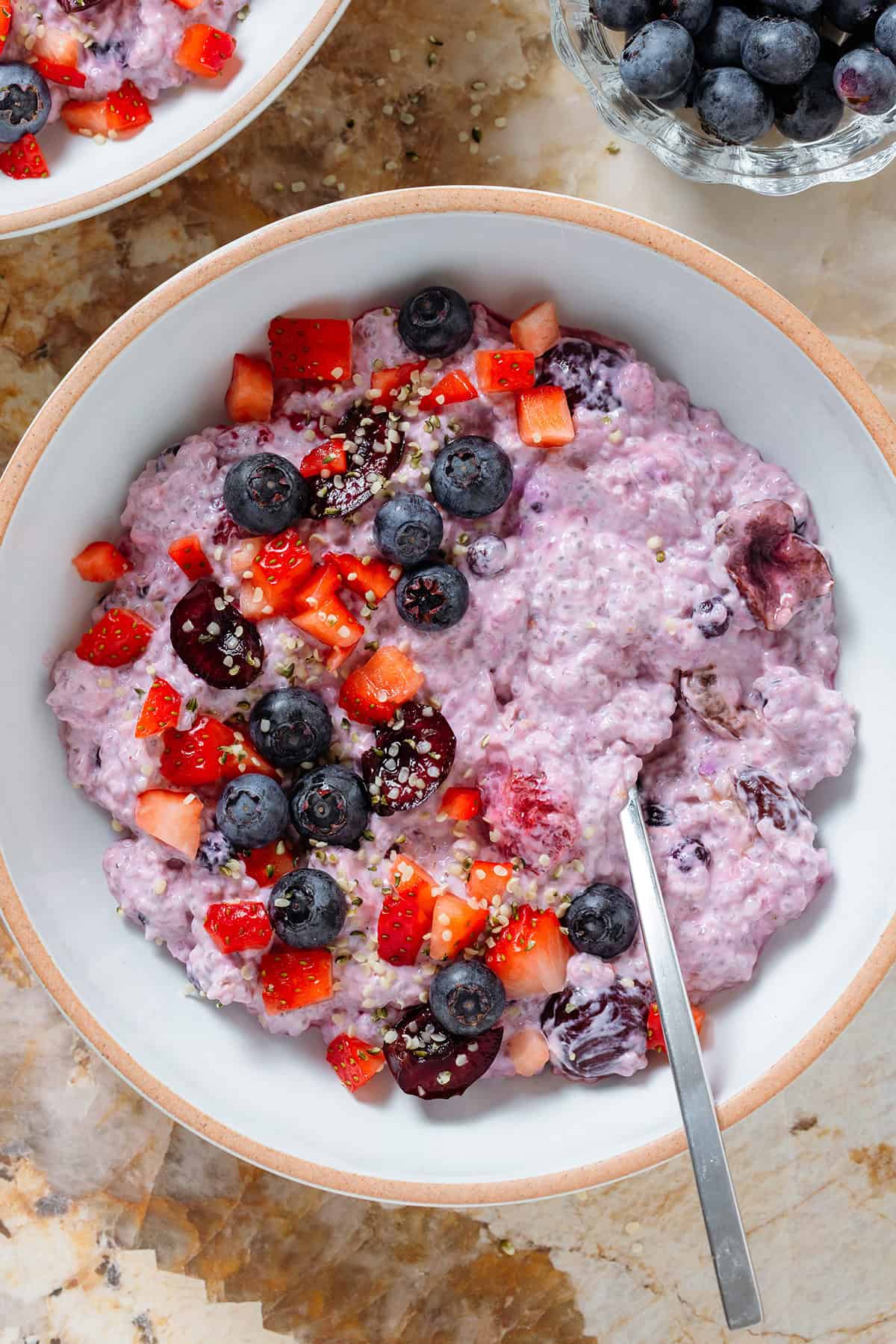 Light purple chia pudding with mixed berries in a low white bowl topped with fresh blueberries, cherries, and chopped strawberries with a spoon in the bowl.