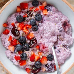 Light purple chia pudding with mixed berries in a low white bowl topped with fresh blueberries, cherries, and chopped strawberries with a spoon in the bowl.