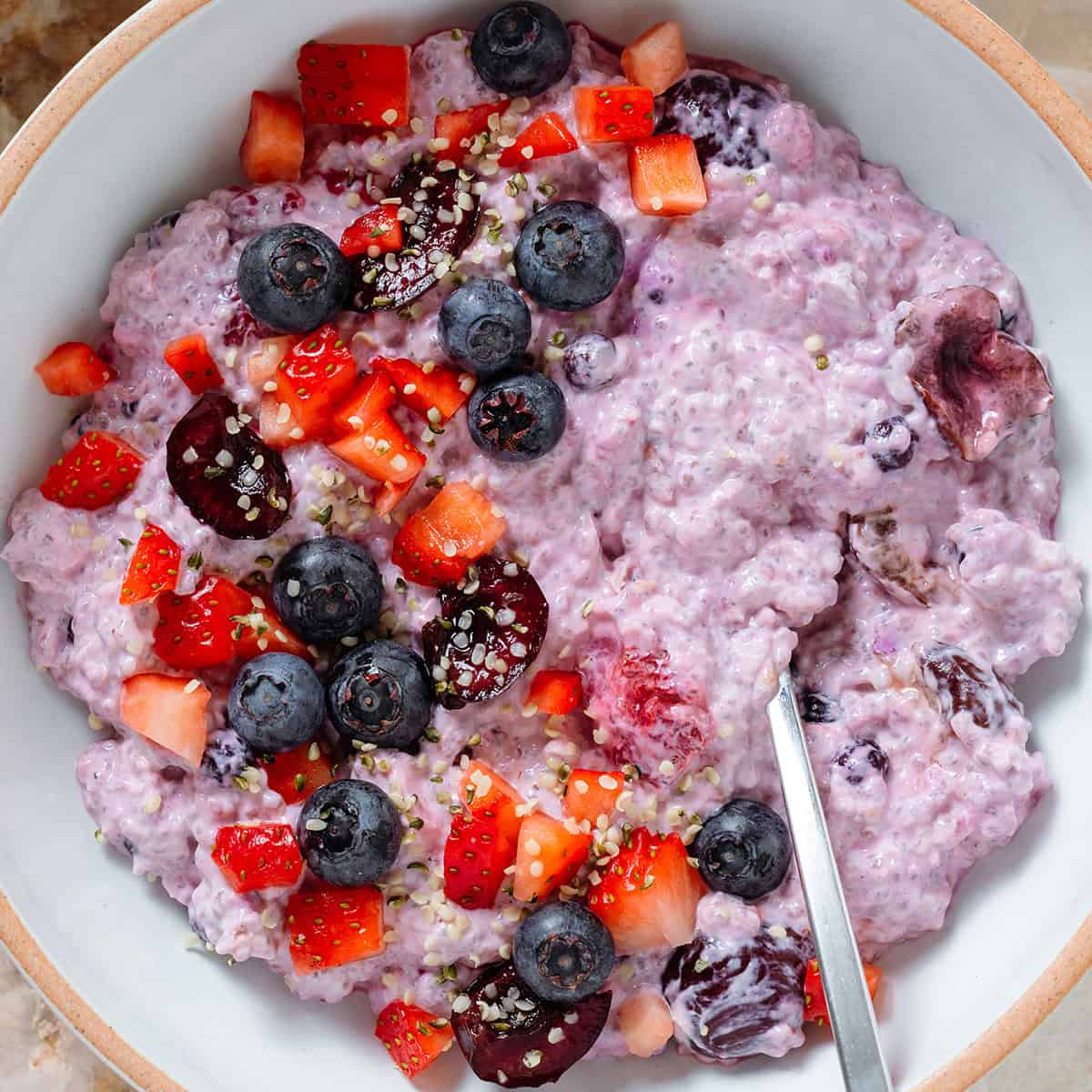 Light purple chia pudding with mixed berries in a low white bowl topped with fresh blueberries, cherries, and chopped strawberries with a spoon in the bowl.