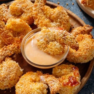 Crispy golden shrimp on a small wooden plate with a creamy dip in the middle and one shrimp dipped into it.