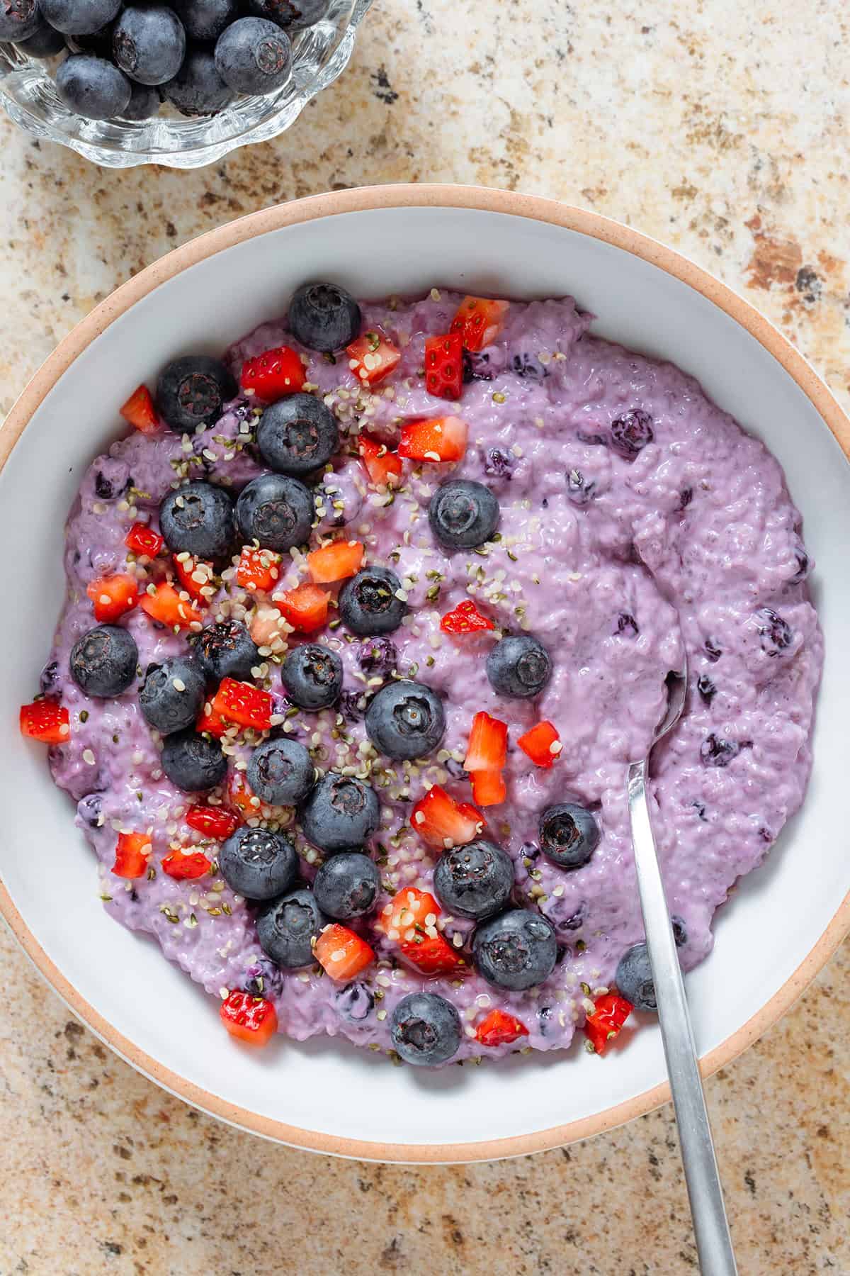 Bright purple blueberry chia pudding in a low white bowl garnished with fresh blueberries and diced strawberries and with a spoon in the bowl.