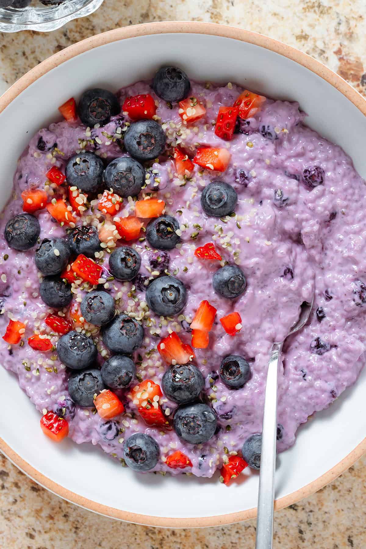 Bright purple blueberry chia pudding in a low white bowl garnished with fresh blueberries and diced strawberries and with a spoon in the bowl.