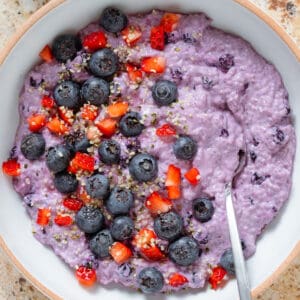 Bright purple blueberry chia pudding in a low white bowl garnished with fresh blueberries and diced strawberries and with a spoon in the bowl.