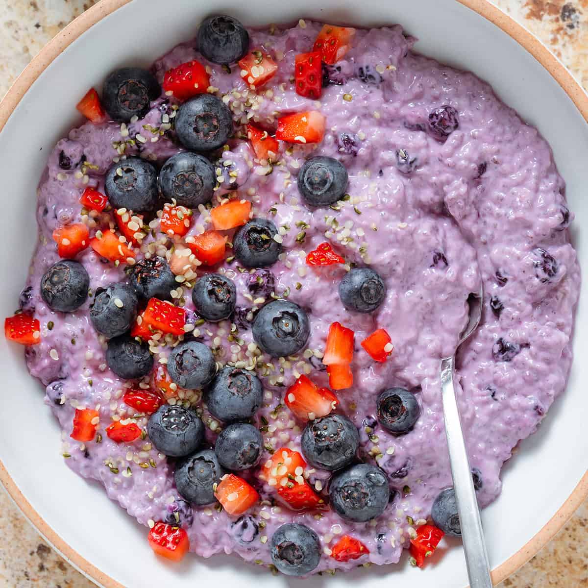 Bright purple blueberry chia pudding in a low white bowl garnished with fresh blueberries and diced strawberries and with a spoon in the bowl.