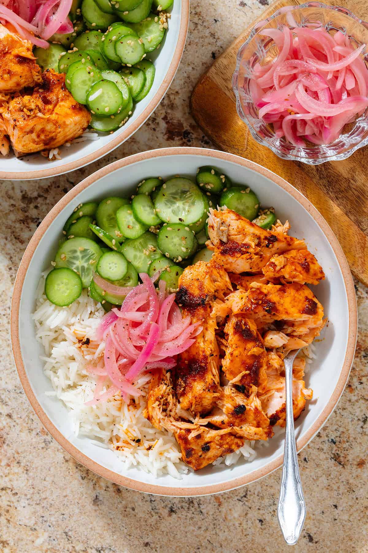 Chili crisp salmon rice bowls with sliced cucumber, sesame seeds, and pickled red onion with a fork in each bowl.