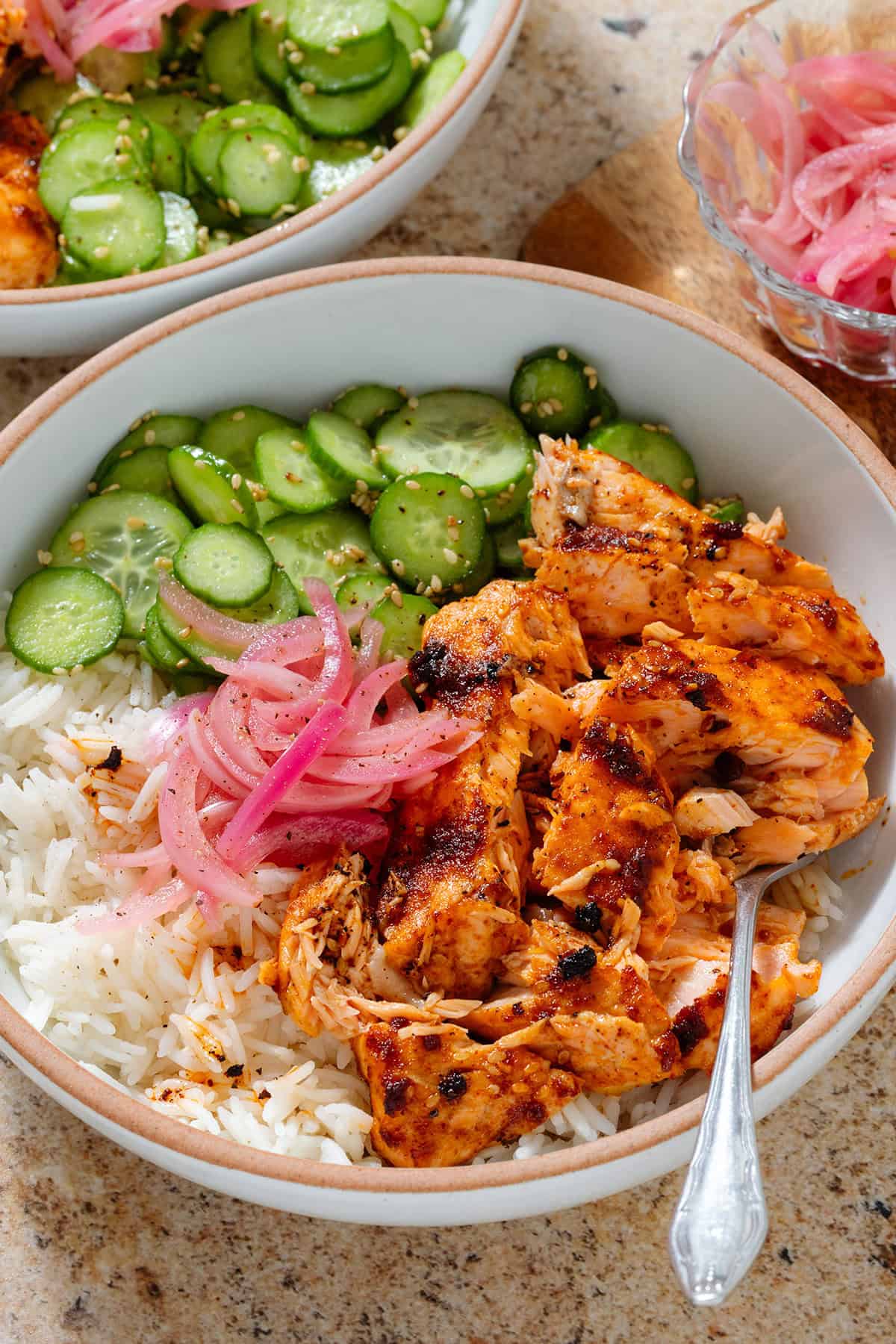 Chili crisp salmon rice bowl with sliced cucumber, sesame seeds, and pickled red onion with a fork in the bowl.
