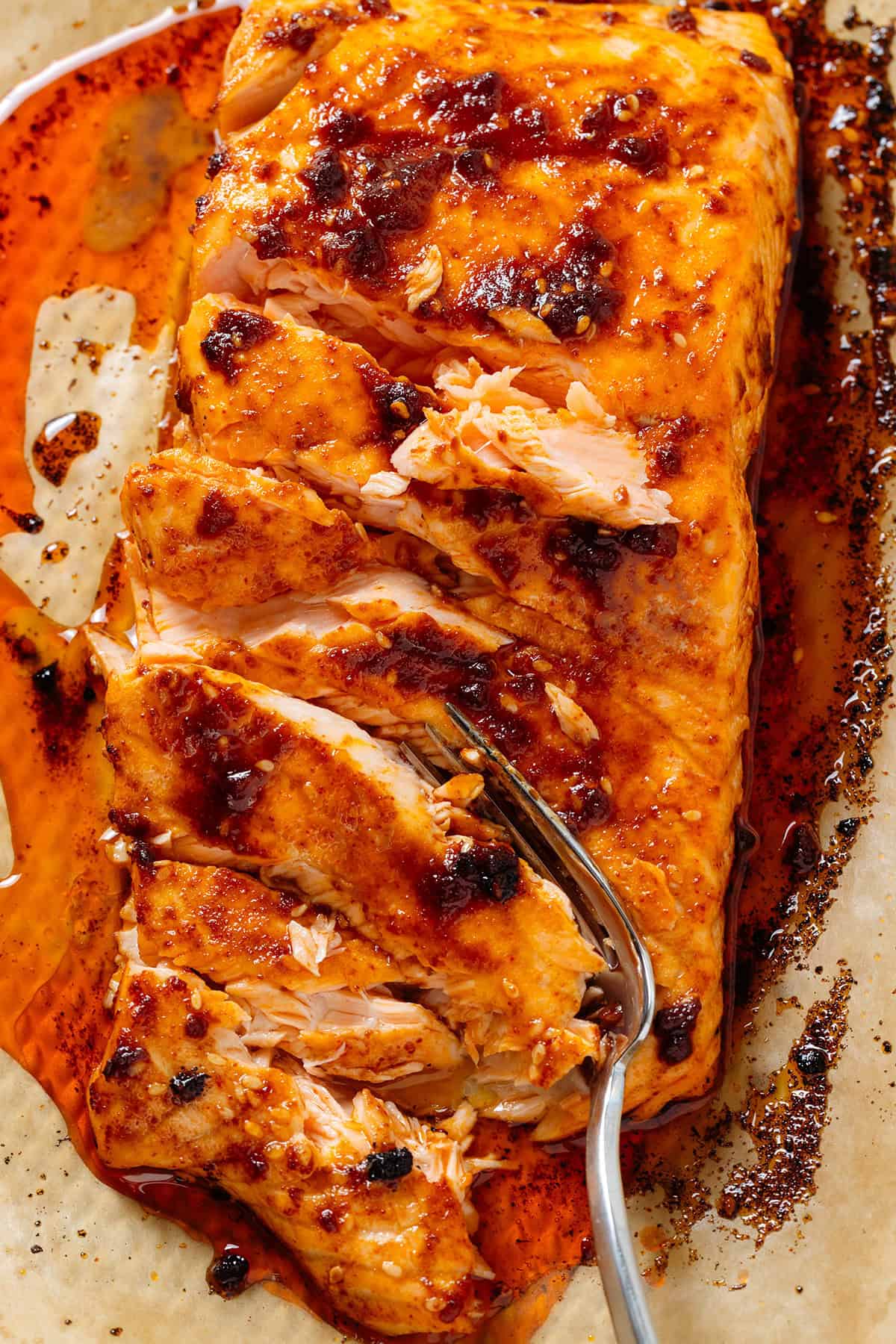 Caramelized roasted chili crisp salmon on a baking sheet lined with parchment paper with a fork slicing it into chunks.
