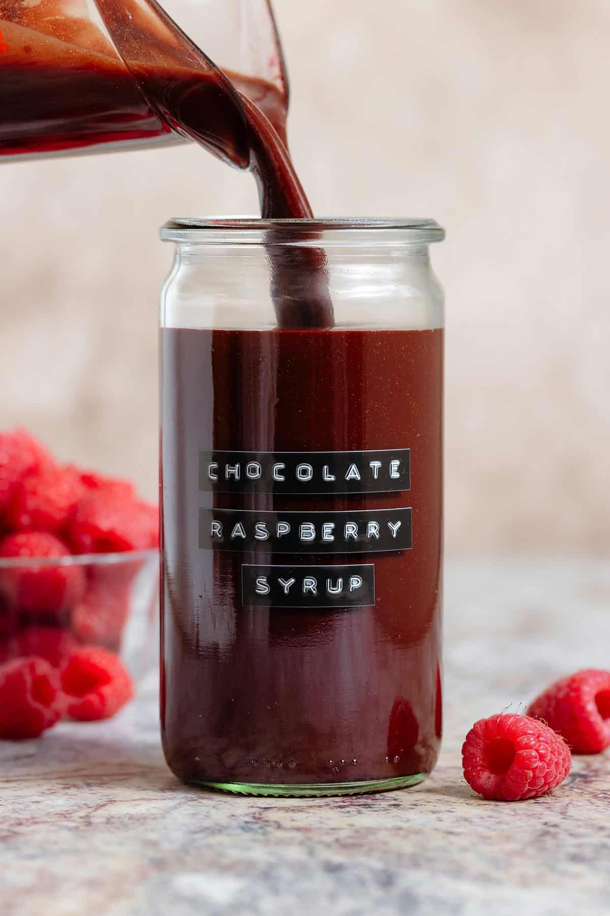 Chocolate raspberry syrup being poured into a tall glass jar with an embossed label and with fresh raspberries all around.