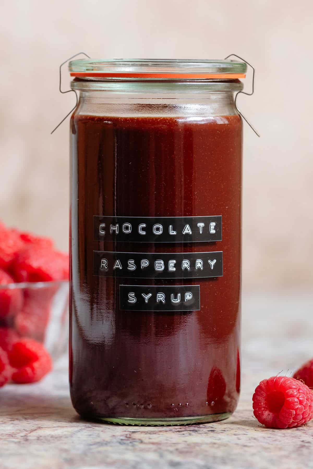 Chocolate raspberry syrup in a tall glass jar with an embossed label and with fresh raspberries all around.