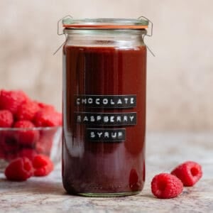 Chocolate raspberry syrup in a tall glass jar with an embossed label and with fresh raspberries all around.