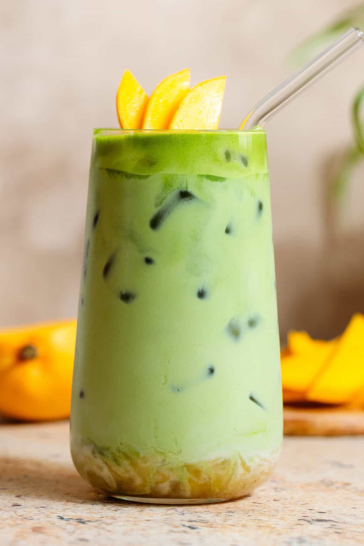 Bright green iced matcha latte in a tall glass with matcha syrup on the bottom, garnished with sliced mango, and with a glass straw.