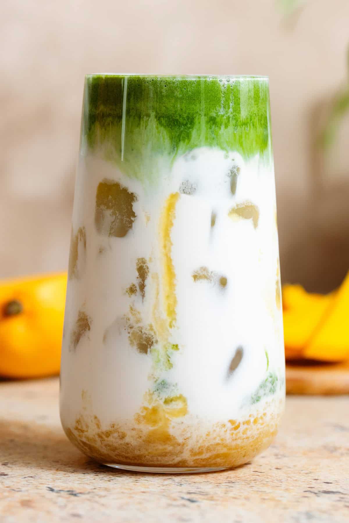 A tall glass with mango syrup, milk, ice, and whisked matcha on top creating a layered look.