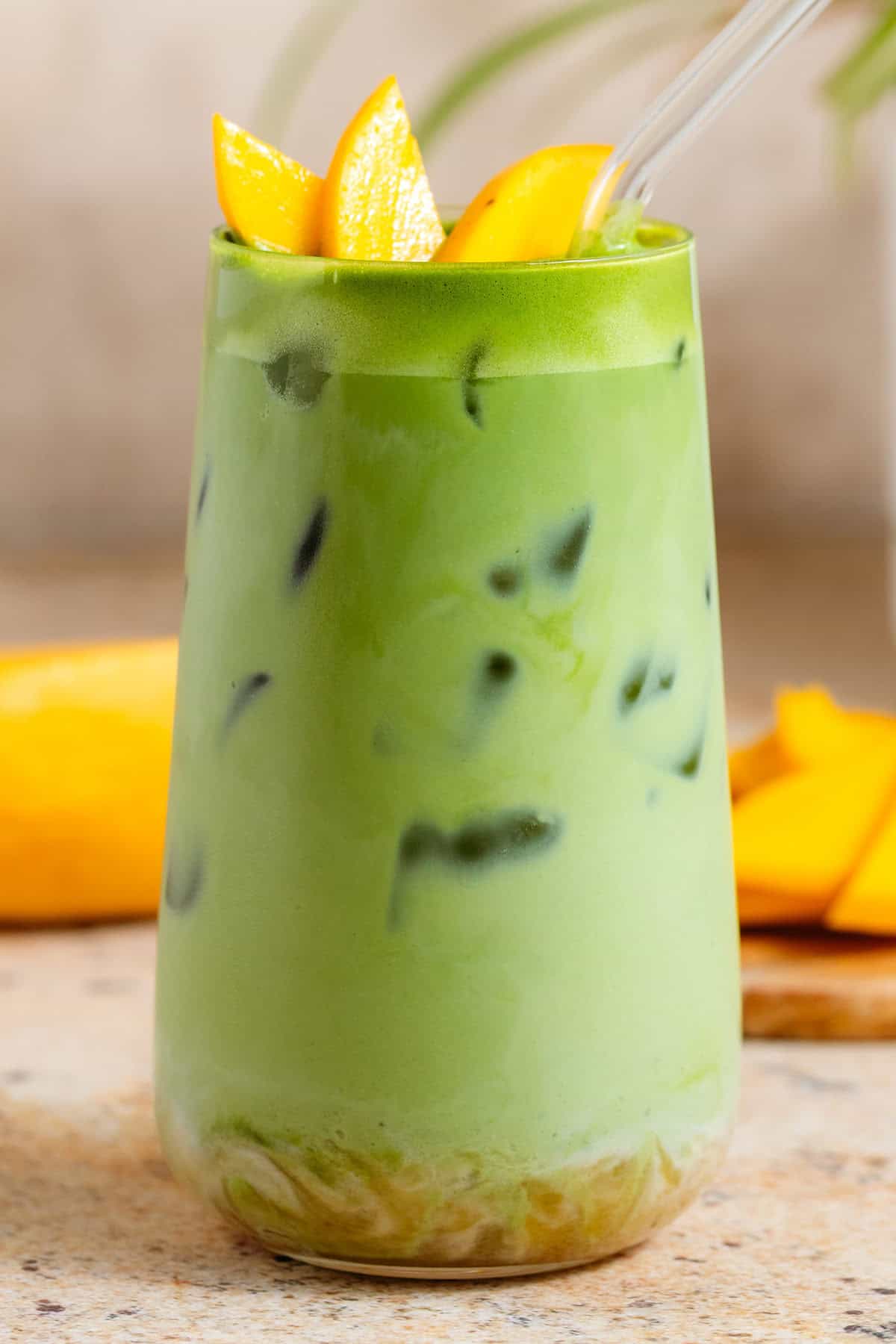 Bright green iced matcha latte in a tall glass with matcha syrup on the bottom, garnished with sliced mango, and with a glass straw.