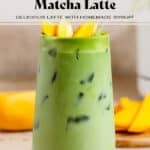 Bright green iced matcha latte in a tall glass with matcha syrup on the bottom, garnished with sliced mango, and with a glass straw.