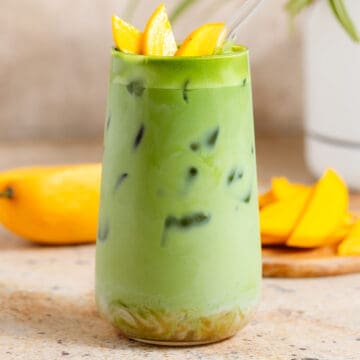 Bright green iced matcha latte in a tall glass with matcha syrup on the bottom, garnished with sliced mango, and with a glass straw.