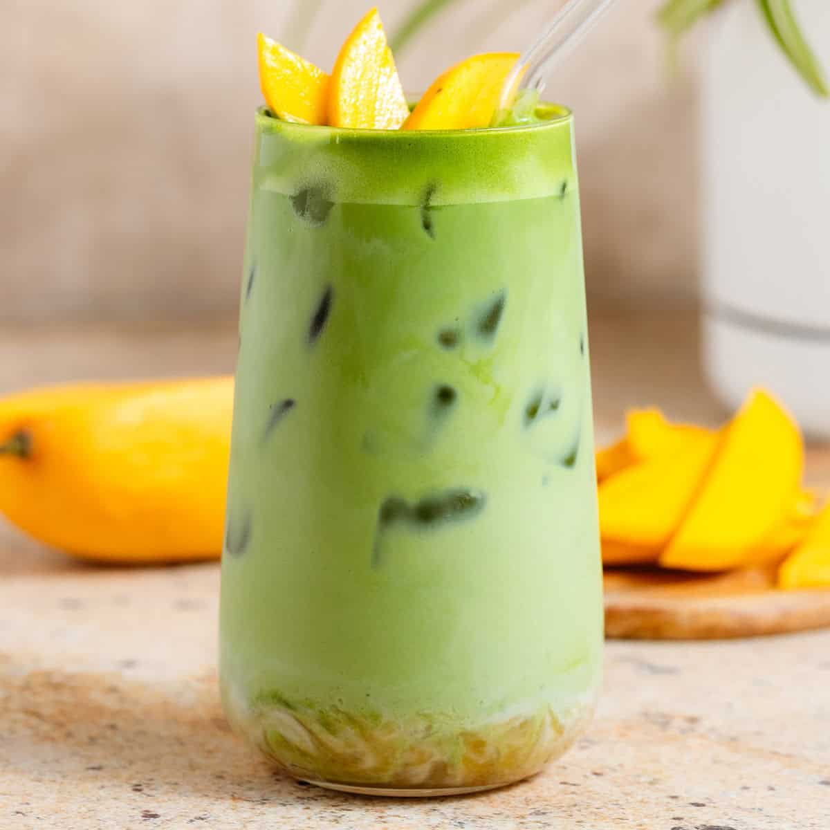 Bright green iced matcha latte in a tall glass with matcha syrup on the bottom, garnished with sliced mango, and with a glass straw.