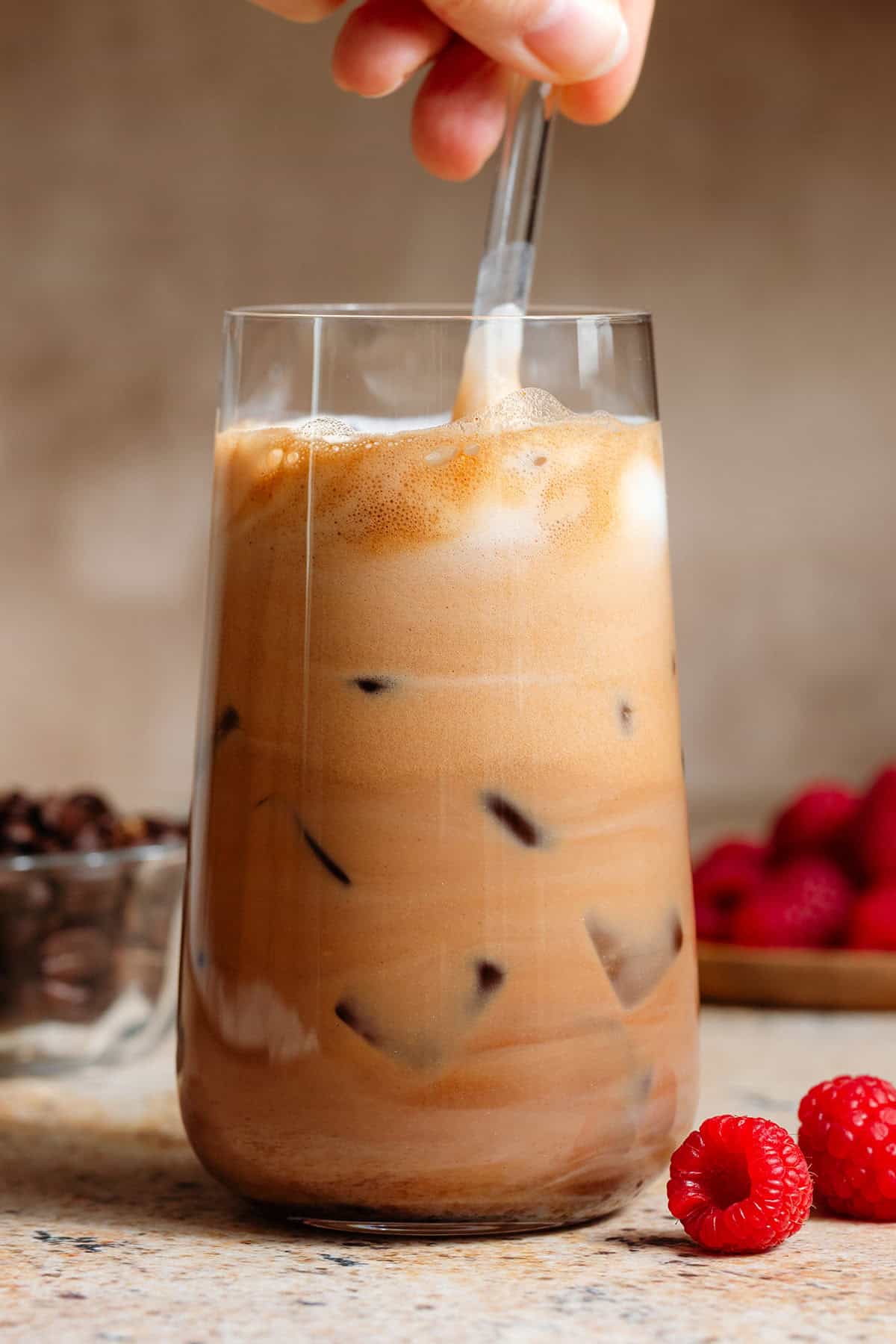 A hand stirring an iced raspberry mocha in a tall glass with a glass straw.