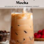 A hand stirring an iced raspberry mocha in a tall glass with a glass straw.