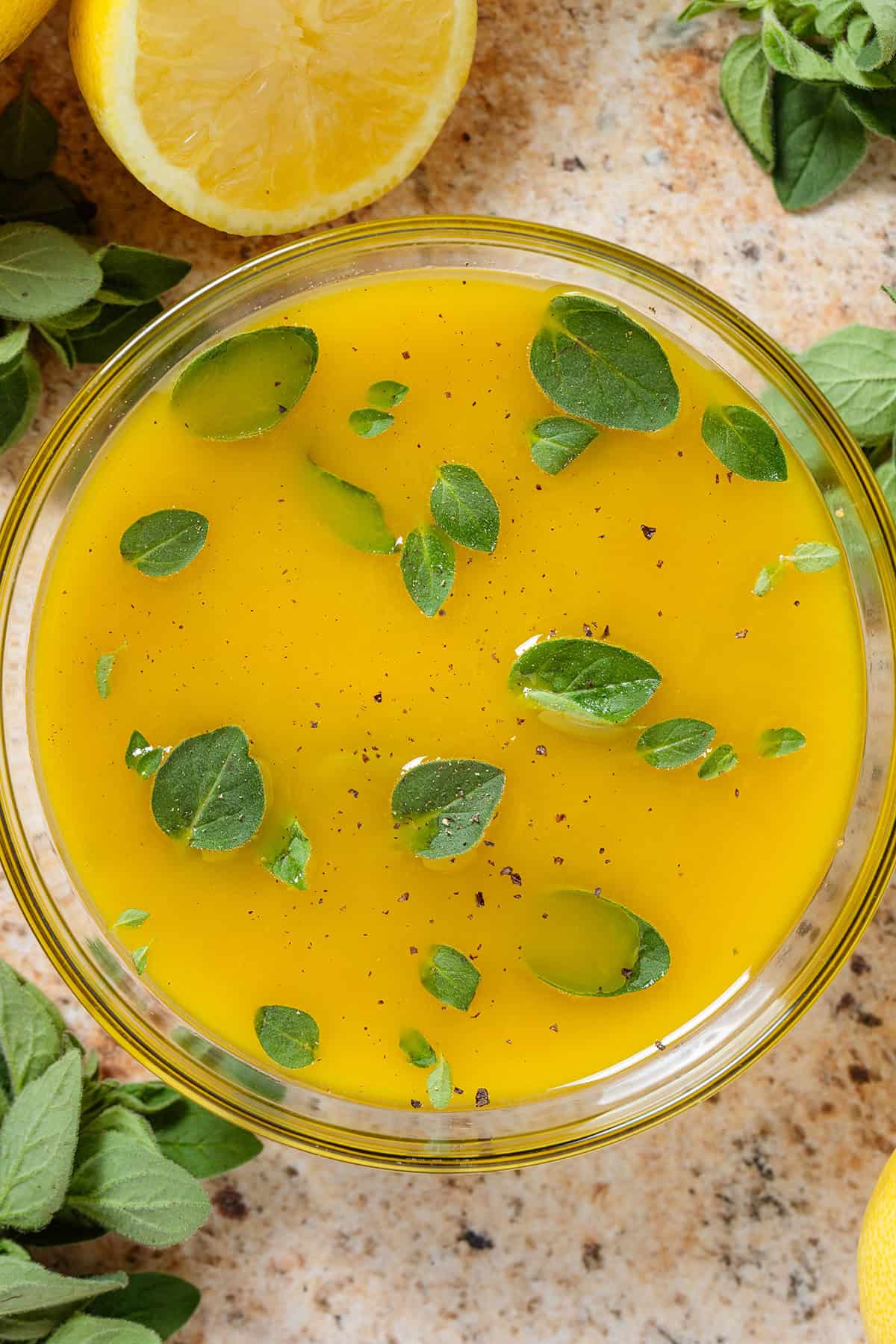 Bright yellow lemon vinaigrette garnished with fresh oregano leaves in a glass bowl with lemons and fresh oregano all around.