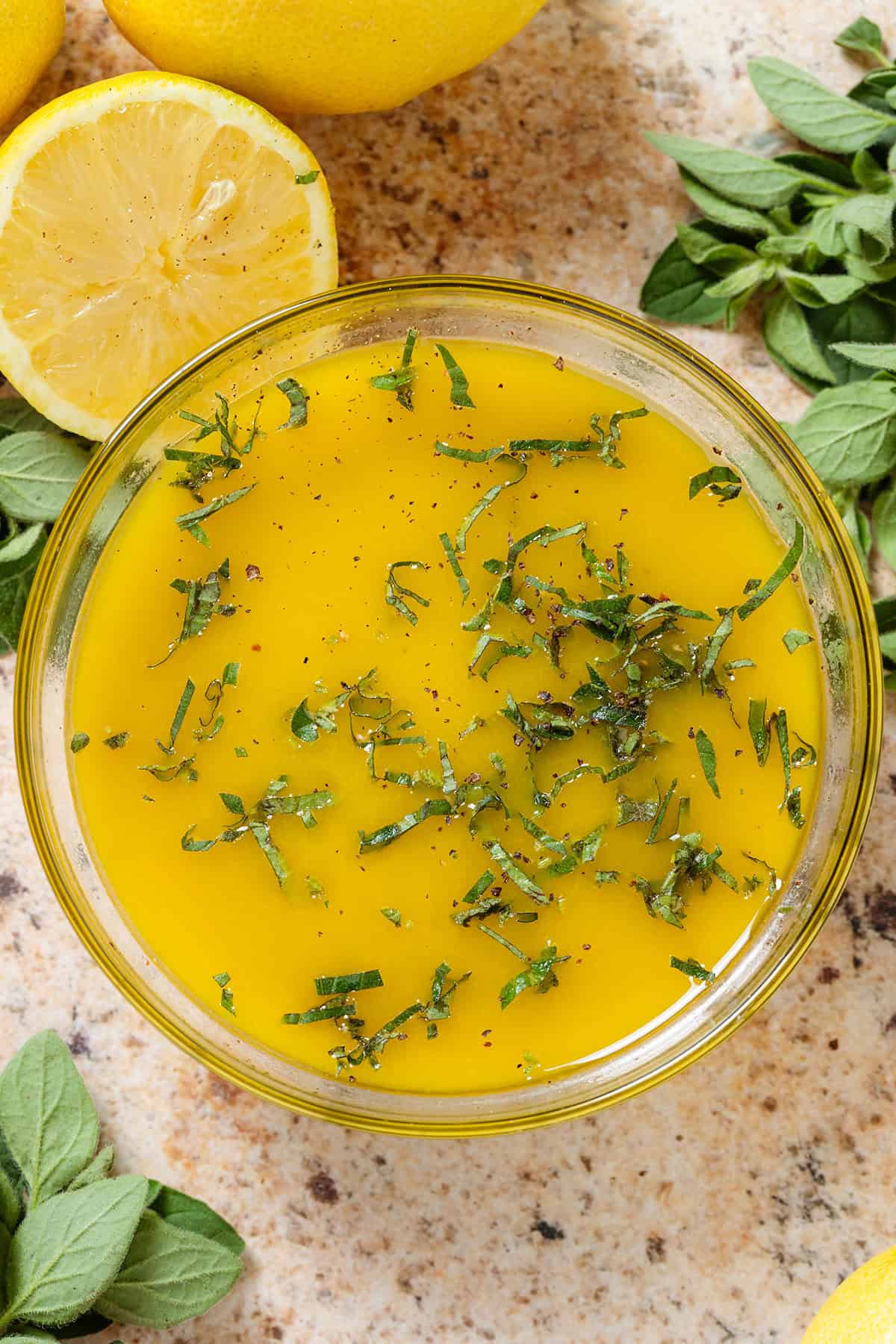 Bright yellow lemon vinaigrette with finely chopped oregano in a glass bowl with more lemons and fresh oregano all around.