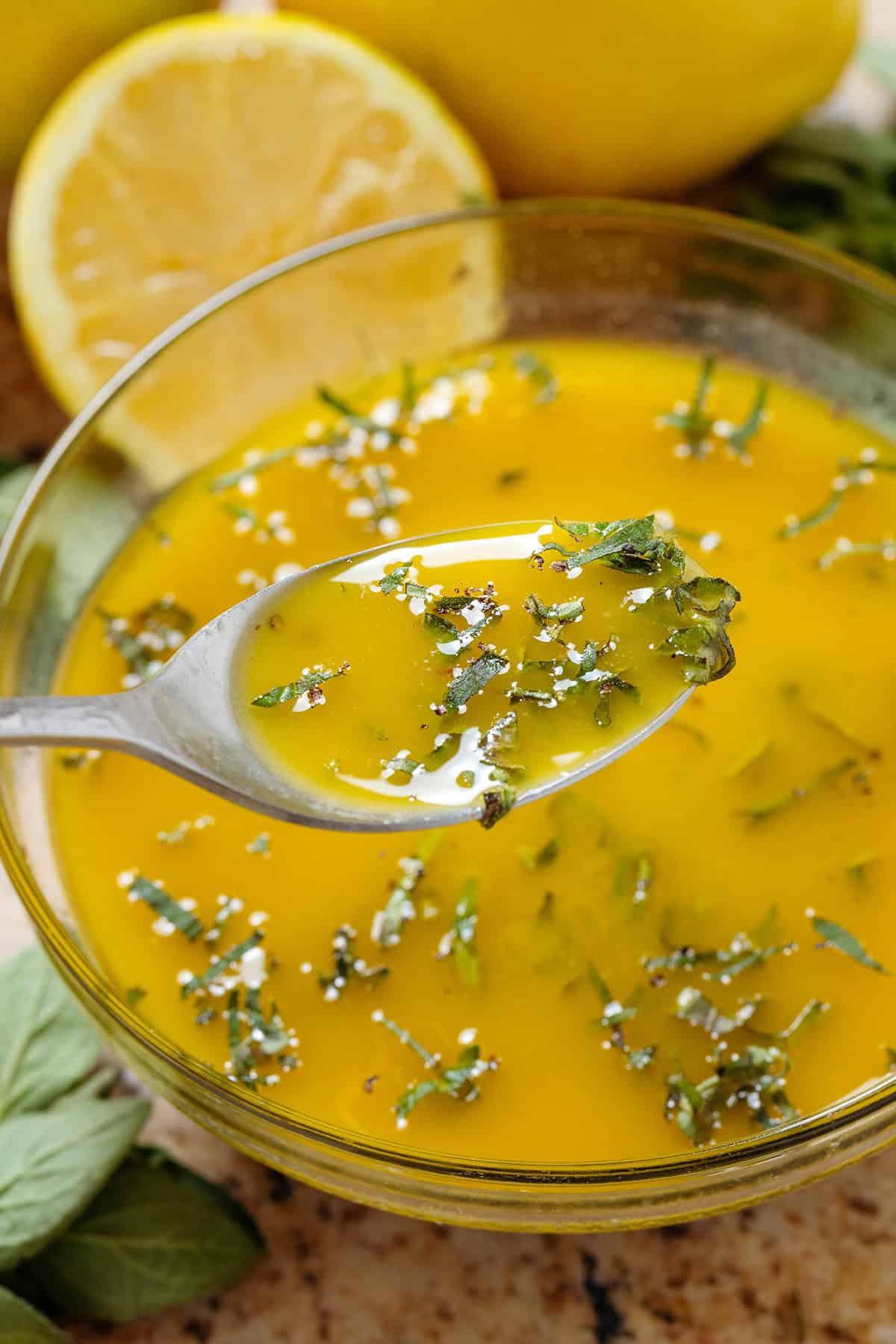 A glass bowl with lemon vinaigrette with finely chopped oregano and a spoon scooping it up.