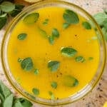 Bright yellow lemon vinaigrette garnished with fresh oregano leaves in a glass bowl with lemons and fresh oregano all around.