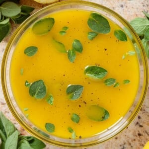 Bright yellow lemon vinaigrette garnished with fresh oregano leaves in a glass bowl with lemons and fresh oregano all around.