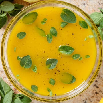 Bright yellow lemon vinaigrette garnished with fresh oregano leaves in a glass bowl with lemons and fresh oregano all around.