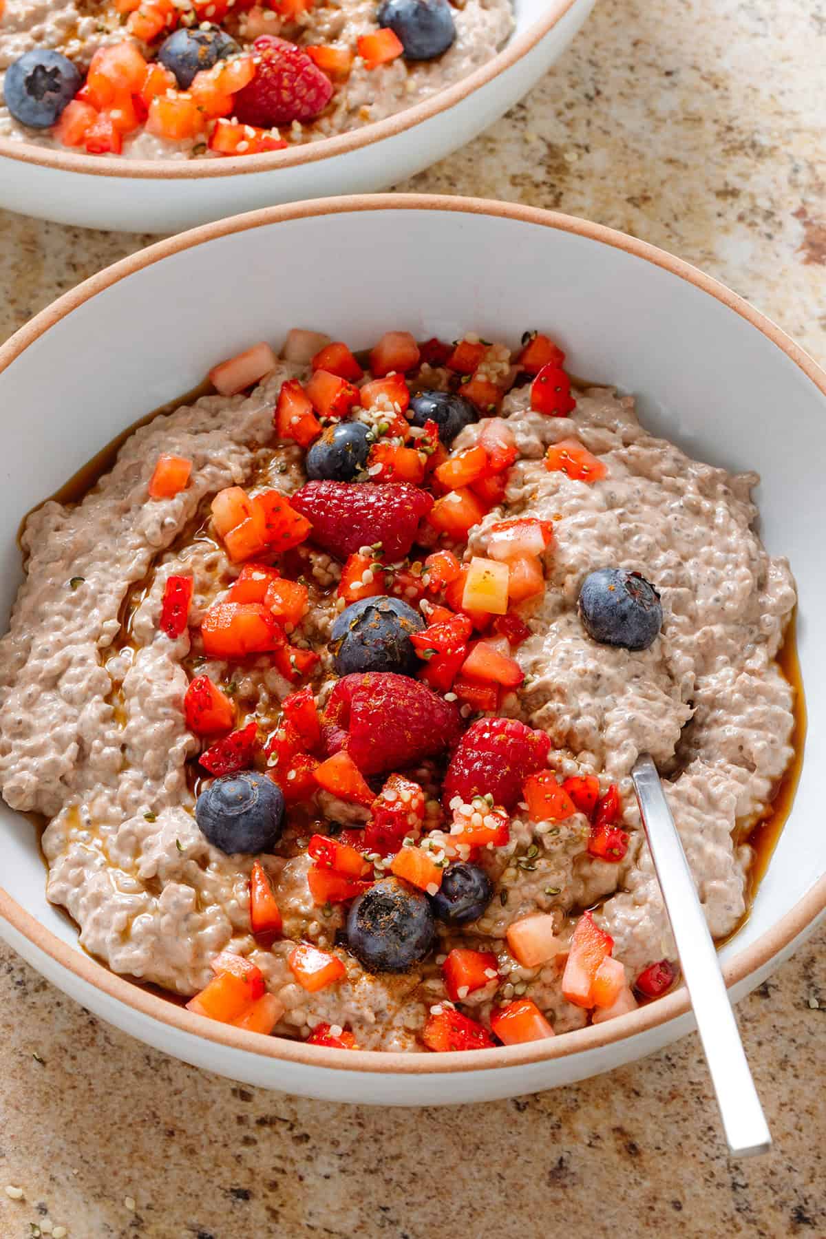 Creamy chocolate protein chia pudding in a white bowl garnished with fresh berries, maple syrup, and seeds with a spoon in the bowl.