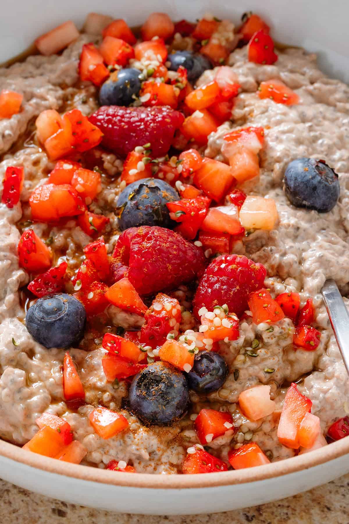 Creamy chocolate protein chia pudding in a white bowl garnished with fresh berries, maple syrup, and seeds with a spoon in the bowl.