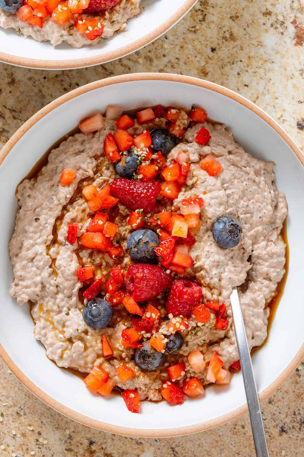 Creamy chocolate protein chia pudding in a white bowl garnished with fresh berries, maple syrup, and seeds with a spoon in the bowl.