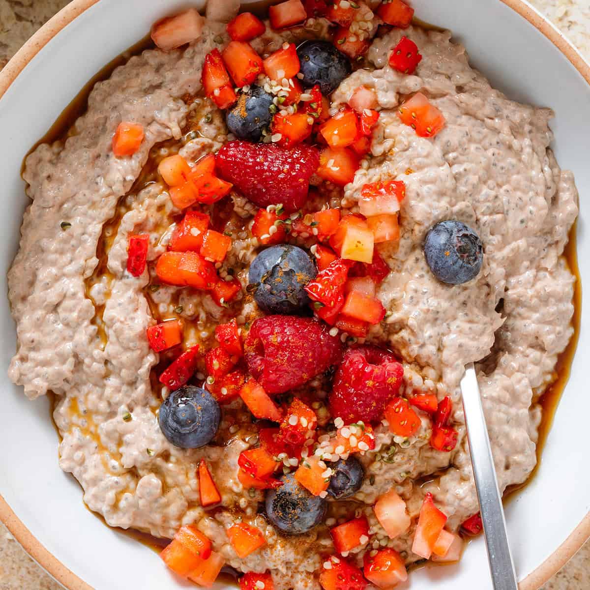 Creamy chocolate protein chia pudding in a white bowl garnished with fresh berries, maple syrup, and seeds with a spoon in the bowl.
