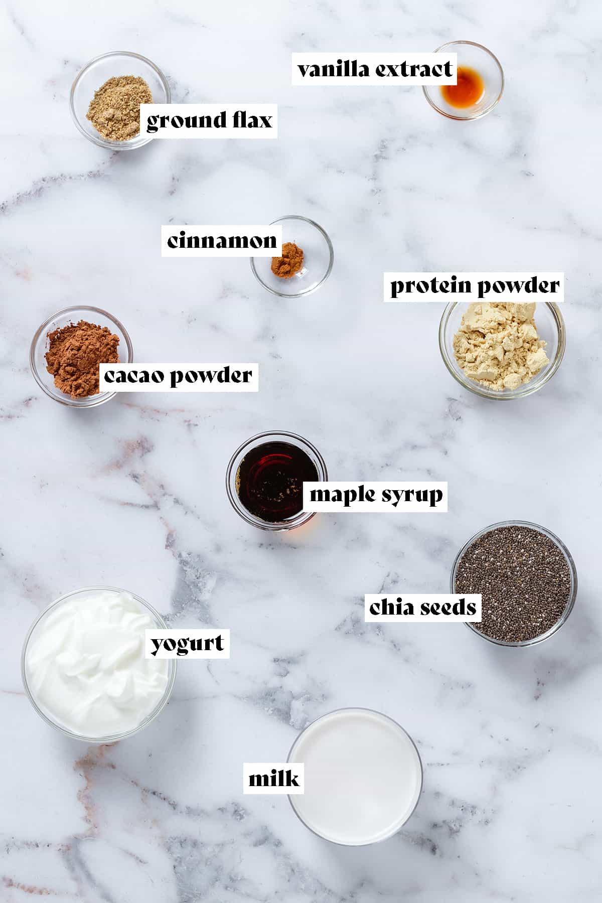 Ingredients in small glass bowls like chia seeds, yogurt, milk, maple syrup, cacao powder, and vanilla.