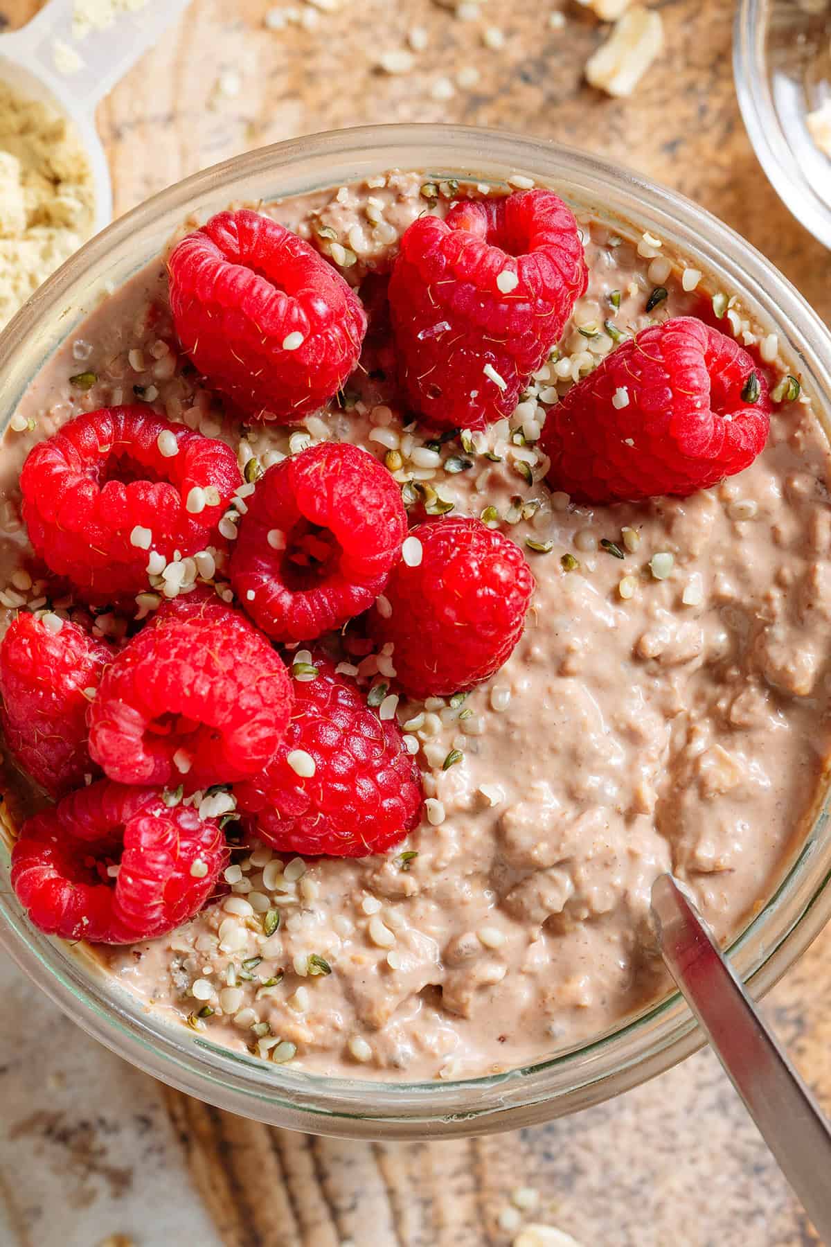 Creamy chocolate protein overnight oats in a glass jar topped with fresh raspberries and seeds with a spoon in the jar.