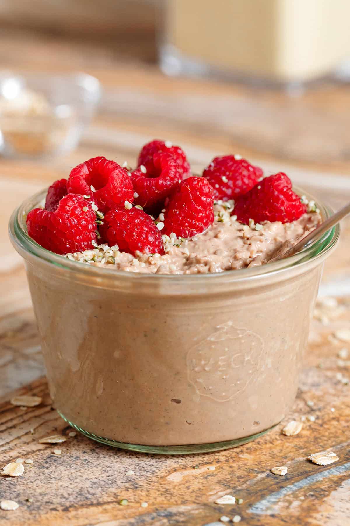Creamy chocolate protein overnight oats in a glass jar topped with fresh raspberries and seeds with a spoon in the jar.