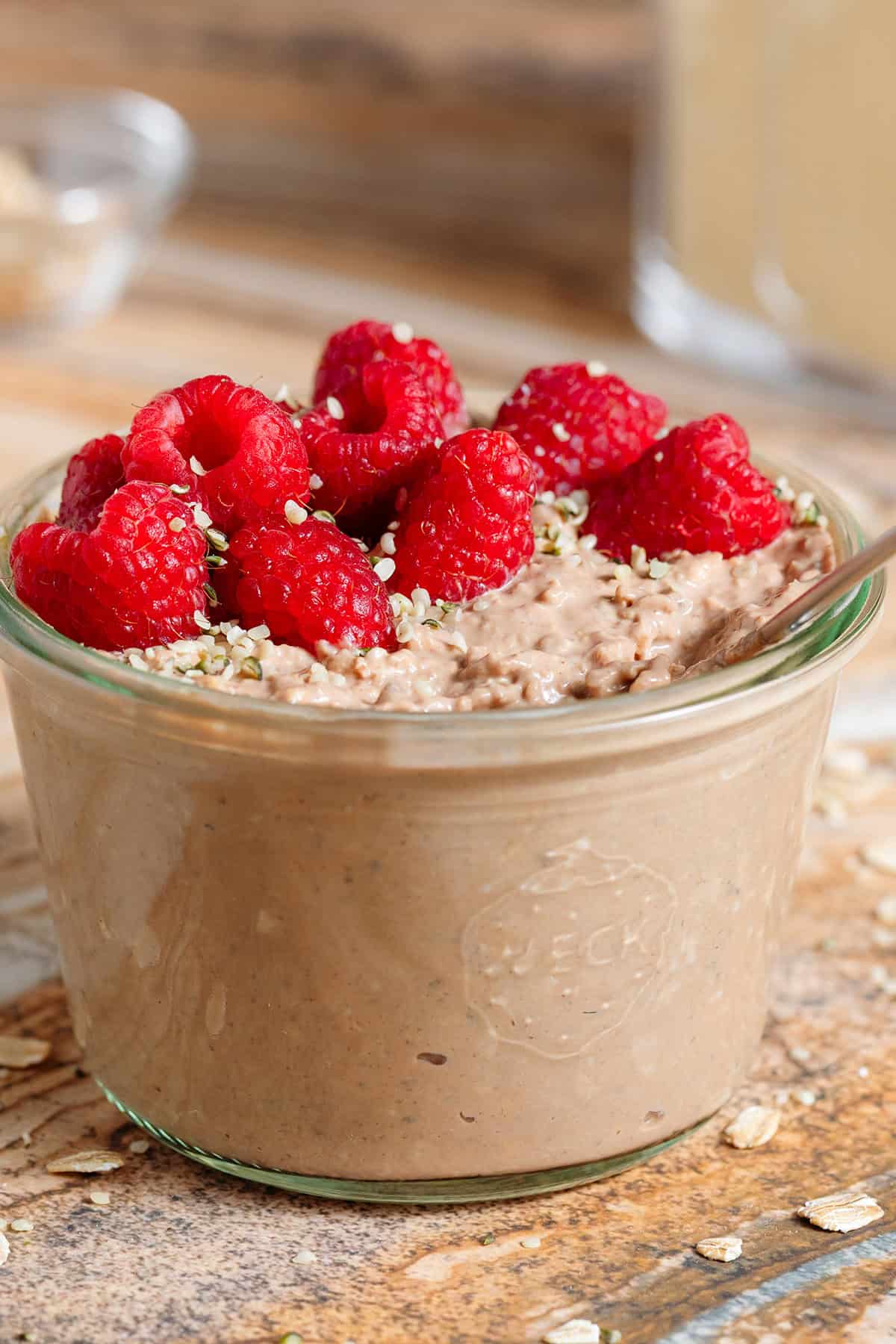 Creamy chocolate protein overnight oats in a glass jar topped with fresh raspberries and seeds with a spoon in the jar.
