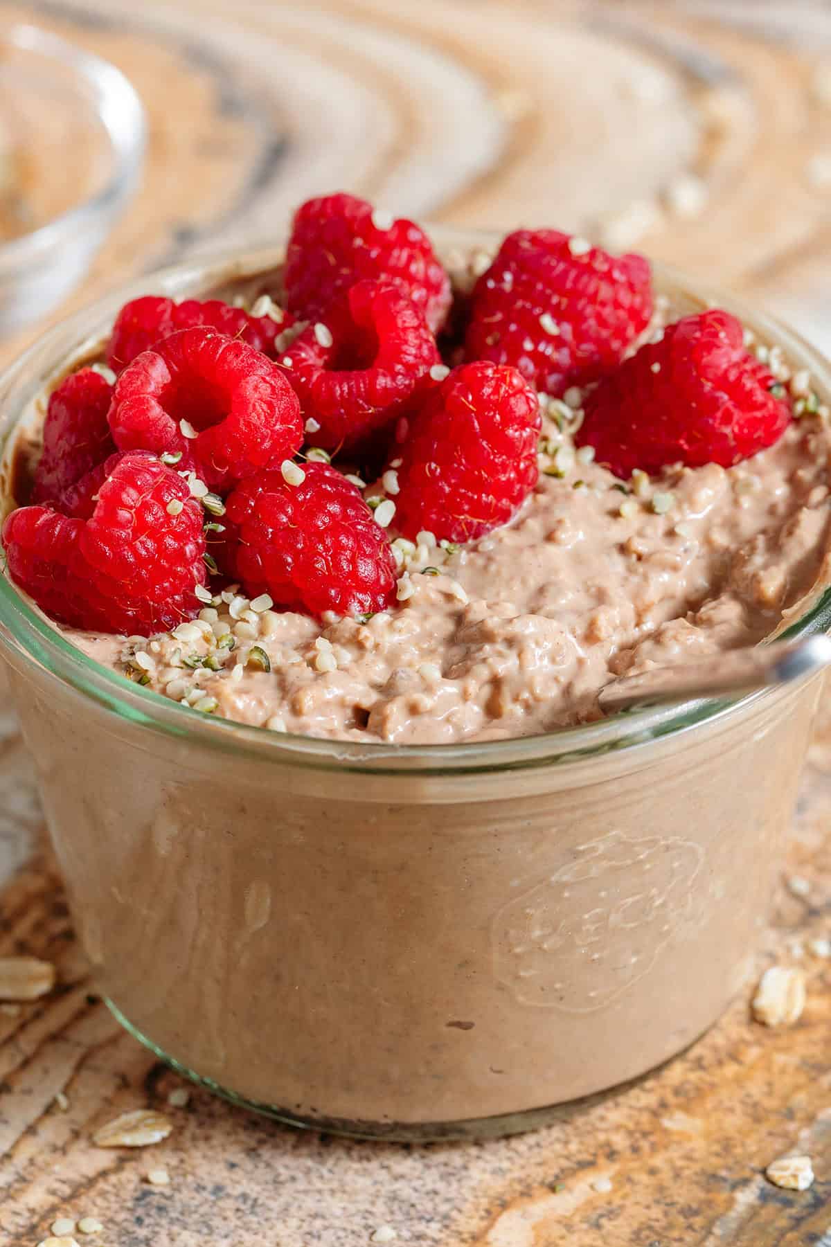 Creamy chocolate protein overnight oats in a glass jar topped with fresh raspberries and seeds with a spoon in the jar.