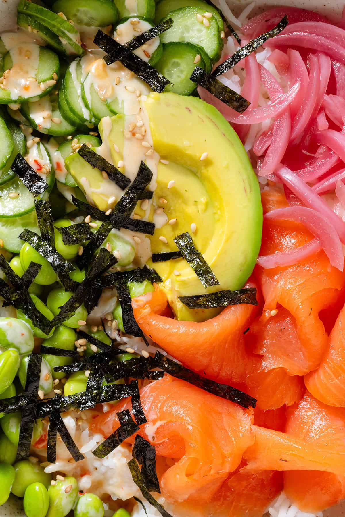 A bowl with smoked salmon, rice, edamame, cucumber, avocado, pickled red onion, strips of nori, spicy mayo, and sesame seeds.