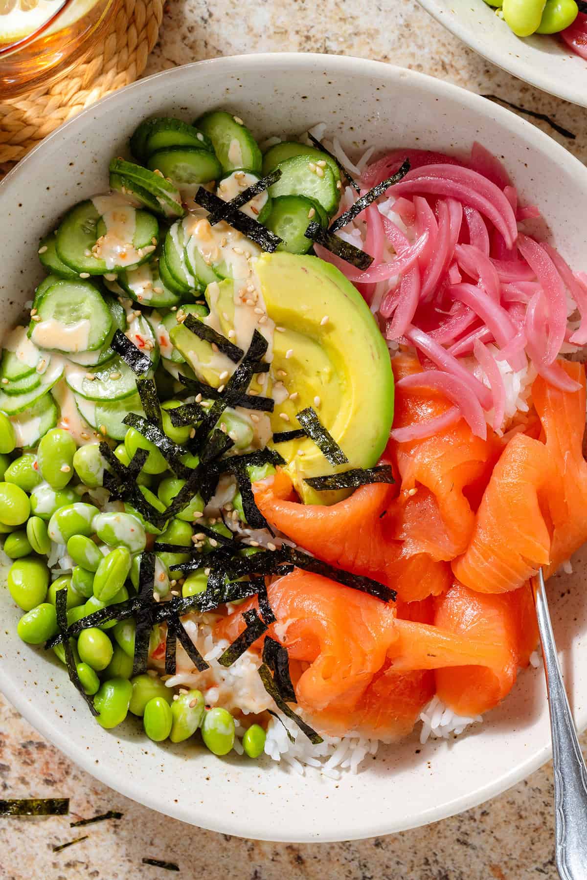 Smoked salmon rice bowl with edamame, cucumber, avocado, pickled red onion, strips of nori, spicy mayo, and sesame seeds.