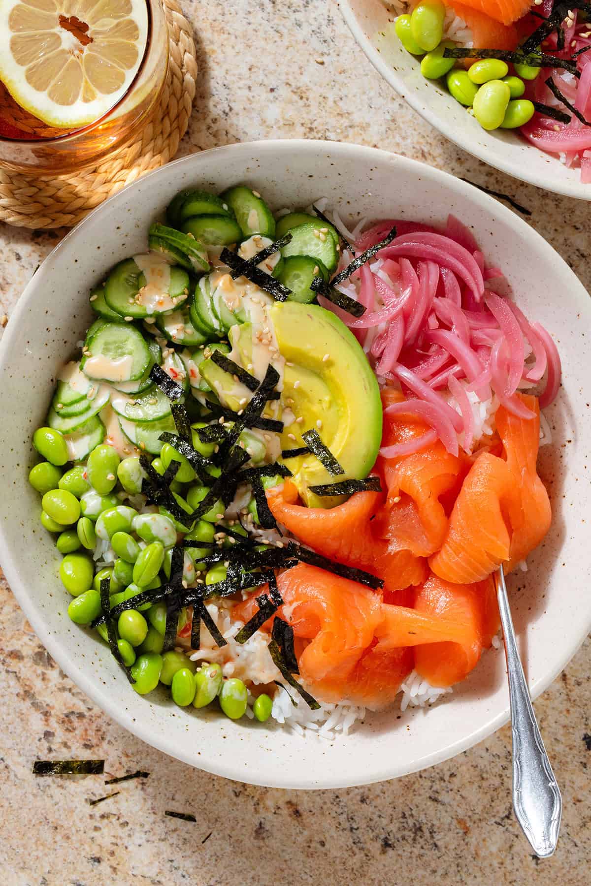 Smoked salmon rice bowl with edamame, cucumber, avocado, pickled red onion, strips of nori, spicy mayo, and sesame seeds.