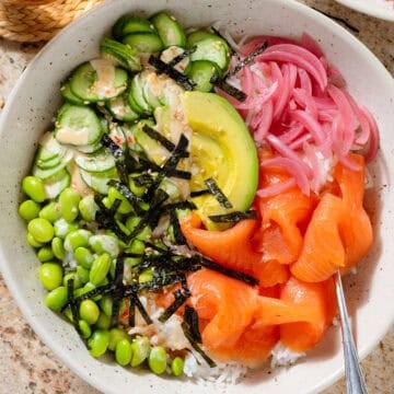 Smoked salmon rice bowl with edamame, cucumber, avocado, pickled red onion, strips of nori, spicy mayo, and sesame seeds.