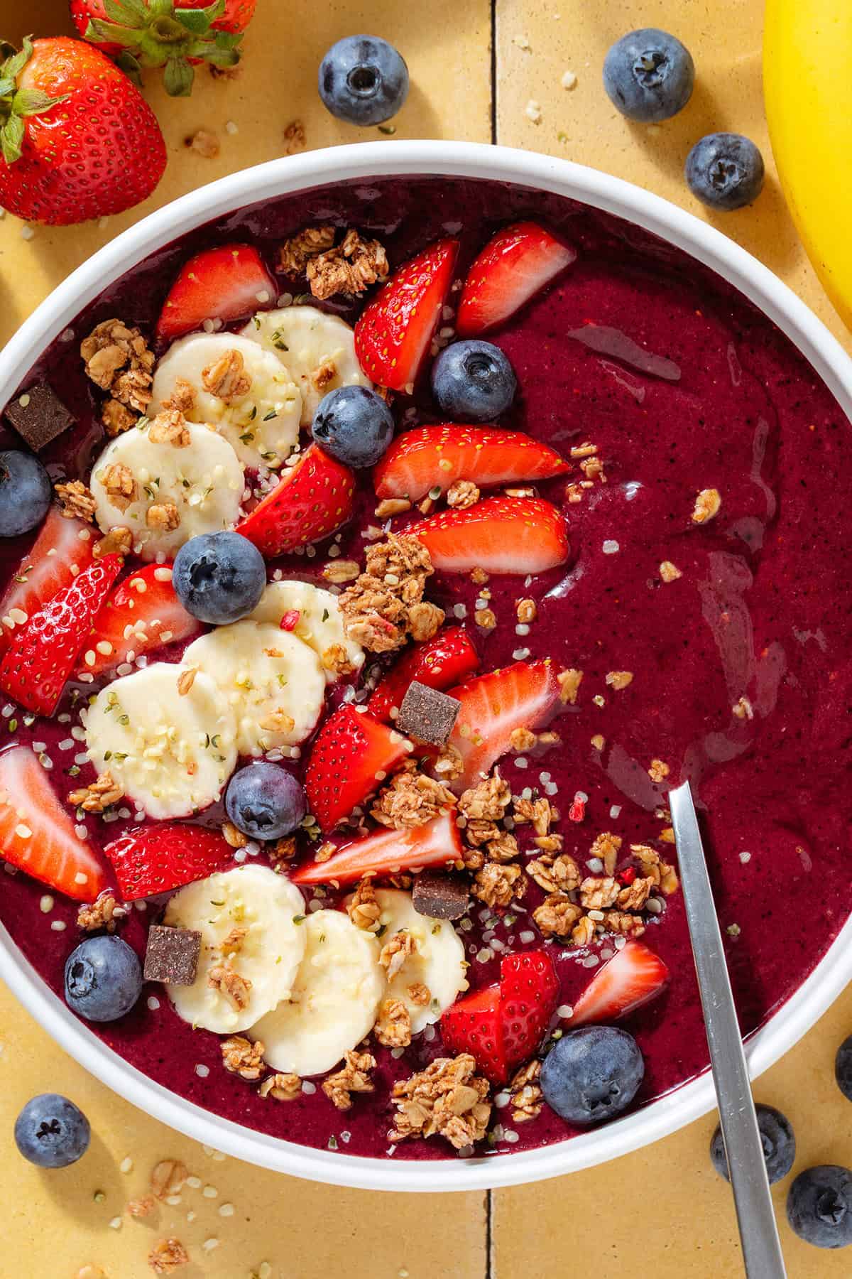 Bright purple acai smoothie in a white bowl topped with sliced banana strawberries, blueberries, and granola, with a spoon in the bowl.