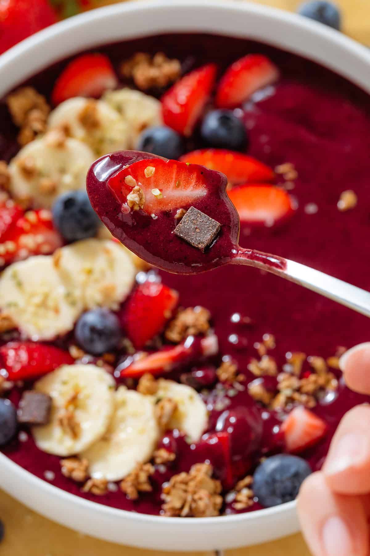 A spoon holding acai smoothie over a white bowl of the smoothie topped with sliced banana strawberries, blueberries, and granola.