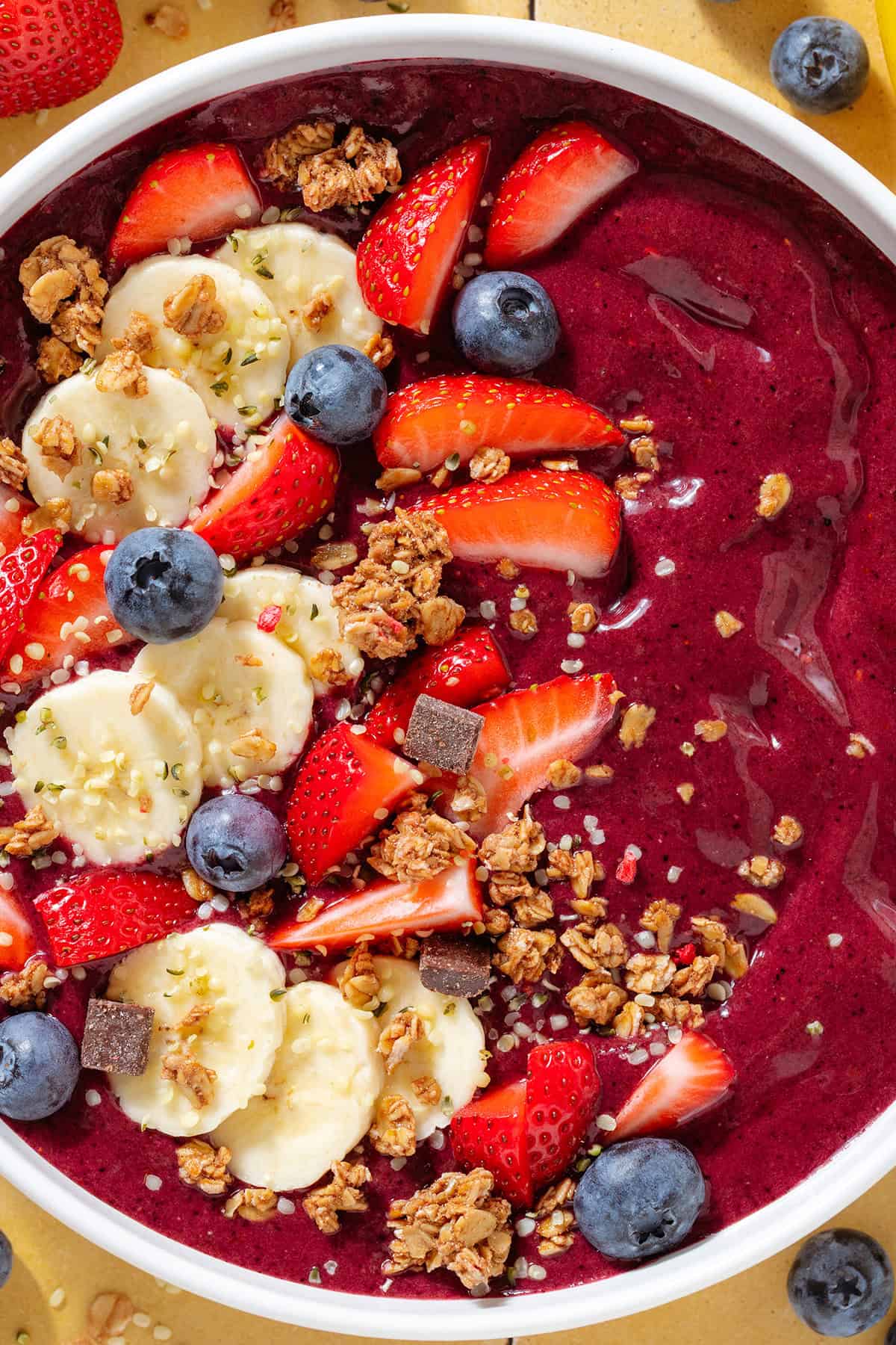 Bright purple acai smoothie in a white bowl topped with sliced banana strawberries, blueberries, and granola.
