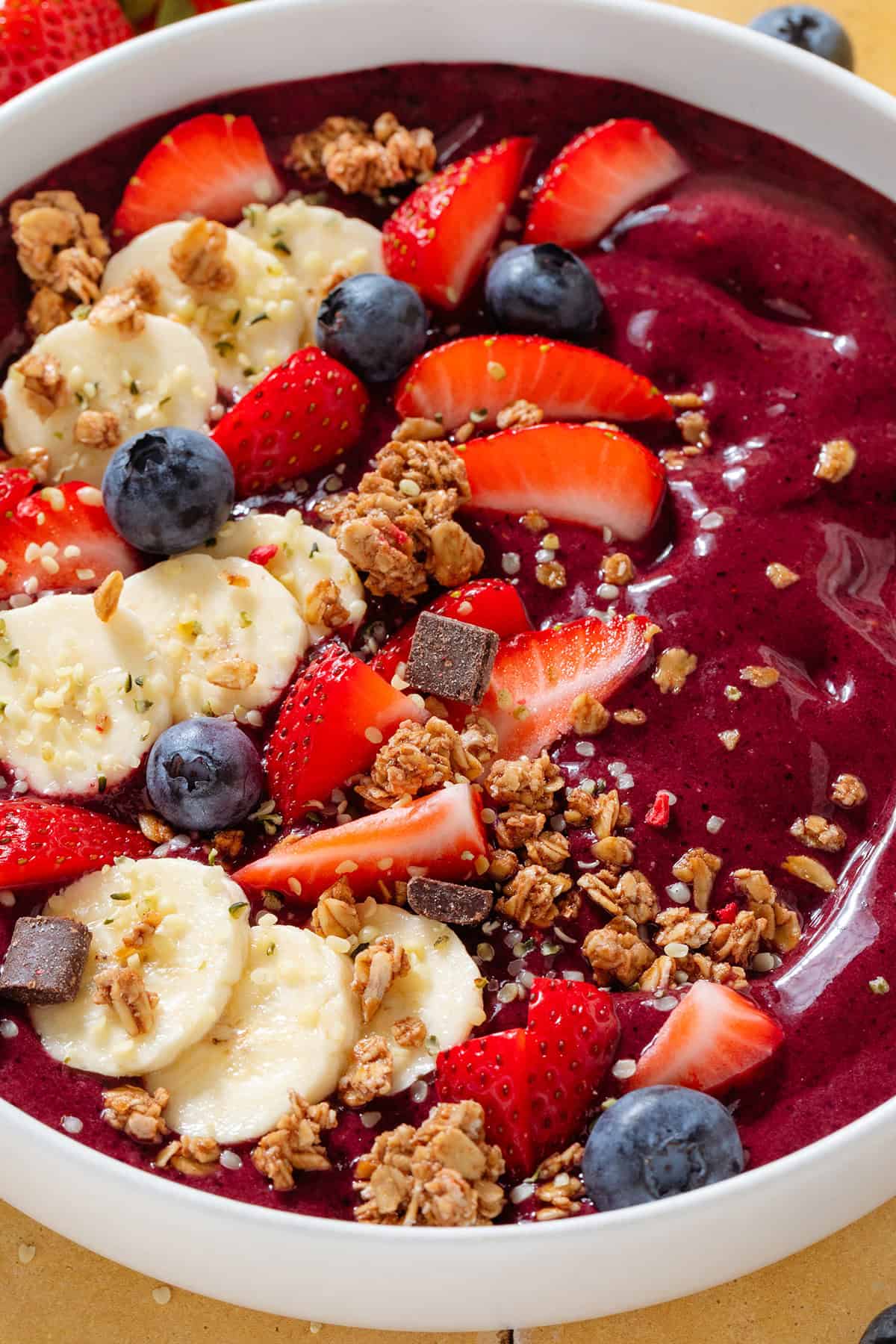 Bright purple acai smoothie in a white bowl topped with sliced banana strawberries, blueberries, and granola.