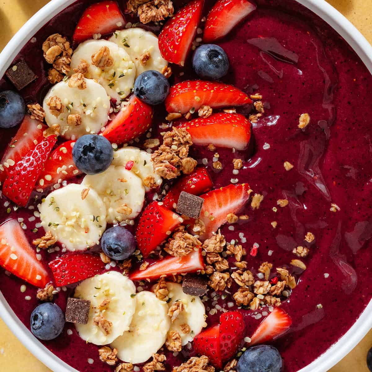 Bright purple acai smoothie in a white bowl topped with sliced banana strawberries, blueberries, and granola.