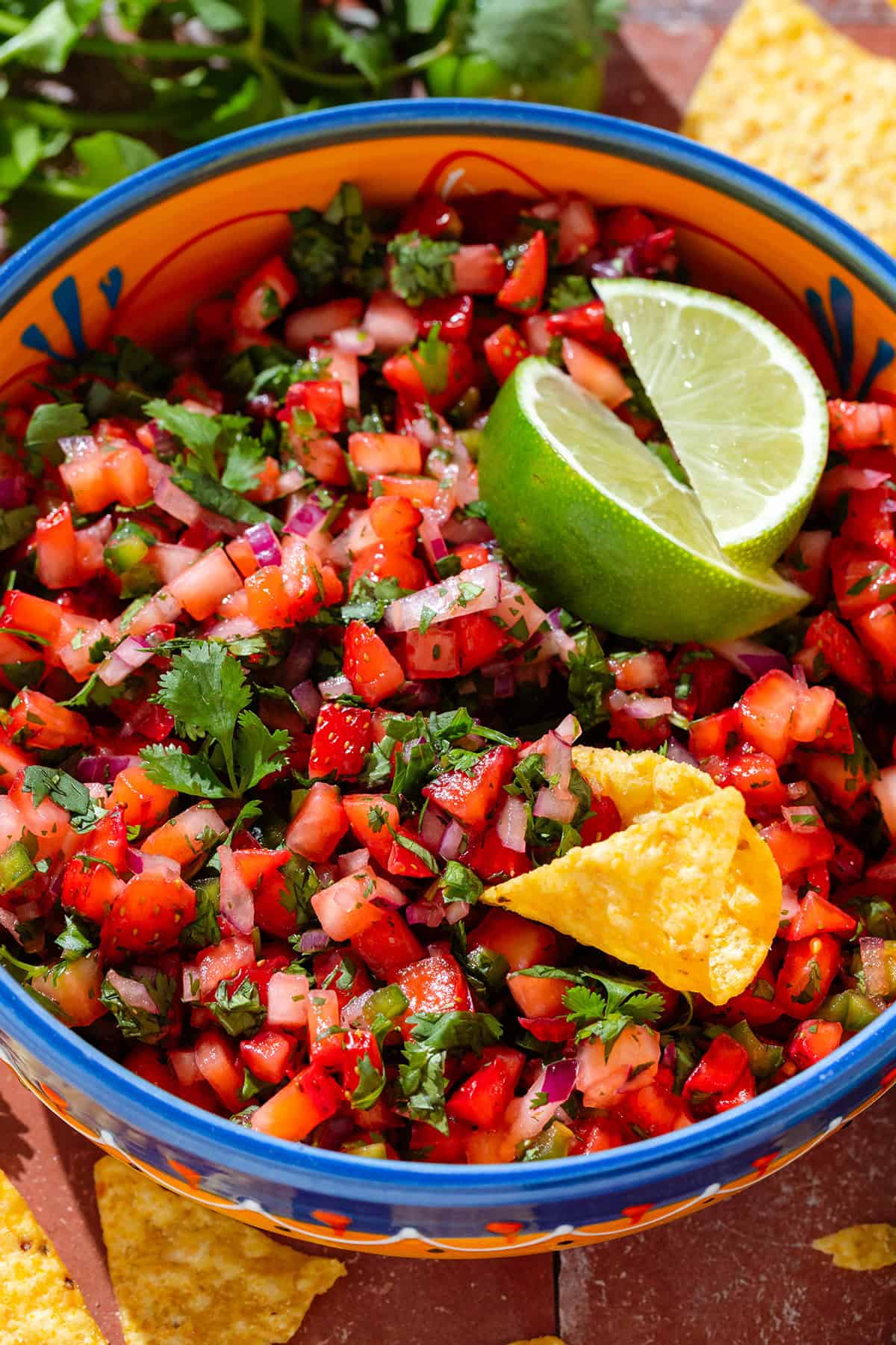 Fresh strawberry salsa in a colorful bowl garnished with two lime wedges and with a tortilla chip dipped into the salsa.