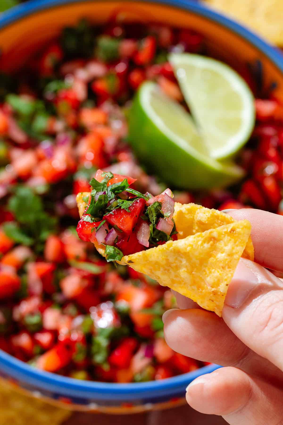 A hand scooping up strawberry salsa using a tortilla chip holding it over the salsa.