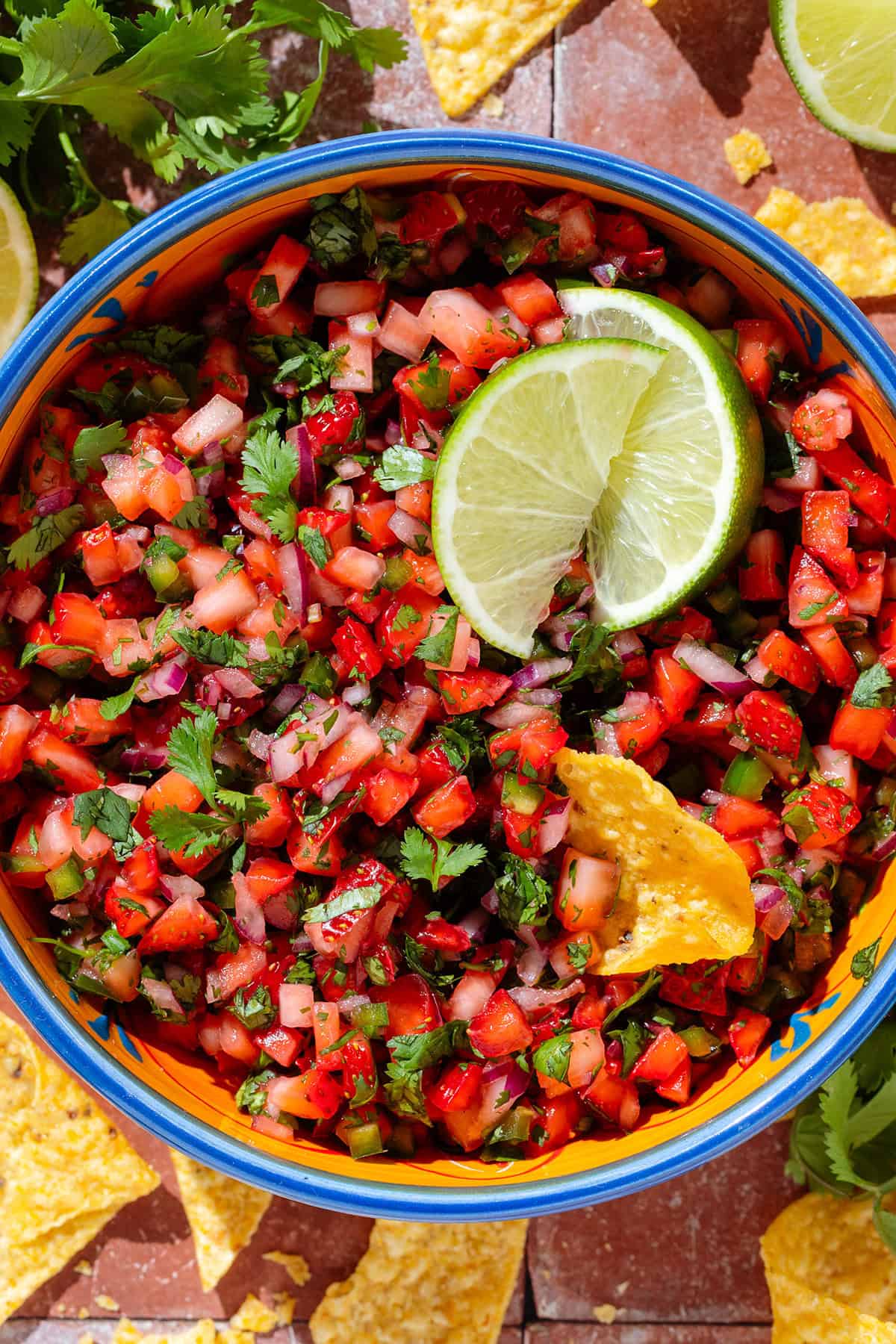 Fresh strawberry salsa in a colorful bowl garnished with two lime wedges and with a tortilla chip dipped into the salsa.