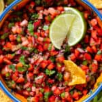 Fresh strawberry salsa in a colorful bowl garnished with two lime wedges and with a tortilla chip dipped into the salsa.